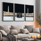 Set of 3 framed canvas triptych showing dramatic night views of Masjid Al-Haram with the Kaaba centered, glowing gold minarets and white marble courtyard against a deep black sky, displayed above a neutral sofa.