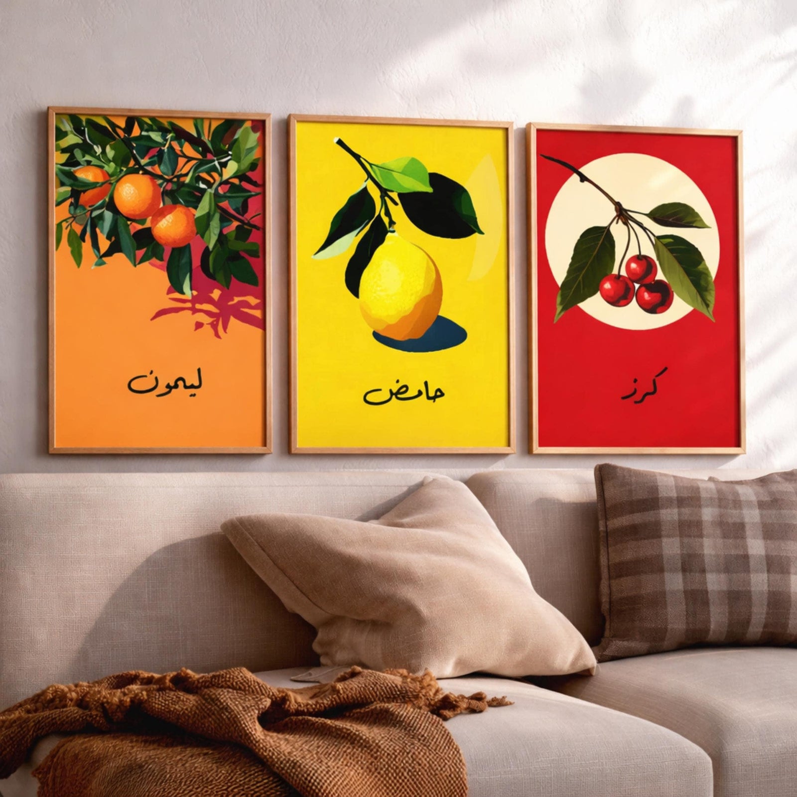 Set of 3 framed canvas triptych featuring bold retro illustrations of orange, lemon and cherry with Arabic calligraphy labels on vibrant orange, yellow and red backgrounds, displayed above a neutral sofa.