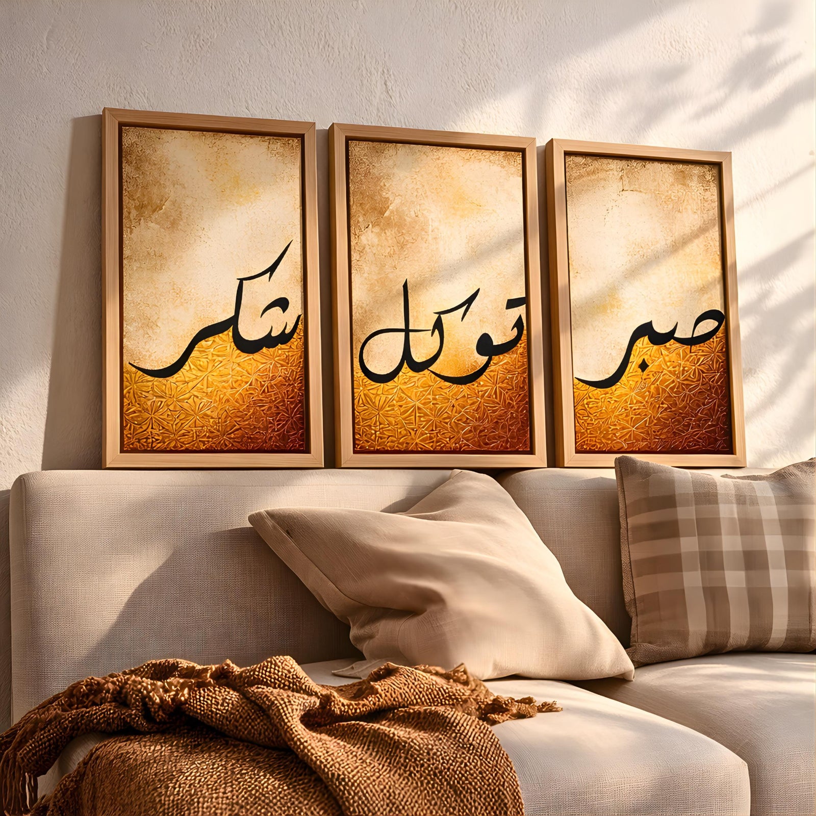 Set of 3 framed canvas triptych featuring Shukr, Tawakkul and Sabr in bold black Arabic calligraphy on a warm amber and cream textured background, displayed above a neutral sofa in a modern living room.