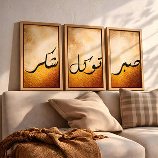 Set of 3 framed canvas triptych featuring Shukr, Tawakkul and Sabr in bold black Arabic calligraphy on a warm amber and cream textured background, displayed above a neutral sofa in a modern living room.
