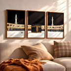 Set of 3 framed canvas triptych showing dramatic night views of Masjid Al-Haram with the Kaaba centered, glowing gold minarets and white marble courtyard against a deep black sky, displayed above a neutral sofa.
