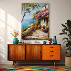 A framed canvas painting depicting a Mediterranean patio with terracotta pots filled with vibrant flowers, a stone tiled floor, and a coastal landscape featuring blue sea and distant mountains under a clear sky, rendered in a realistic style capturing the charm of an Italian seaside terrace.