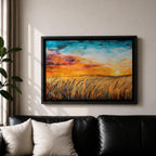A framed canvas painting depicting a wheat field at sunset with golden stalks swaying under a vibrant sky blending orange, yellow, and blue hues. The artwork captures a rustic farmhouse landscape scene with textured brushstrokes emphasizing natural elements and an expansive horizon.