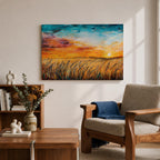 A framed canvas painting depicting a wheat field at sunset with golden stalks swaying under a vibrant sky blending orange, yellow, and blue hues. The artwork captures a rustic farmhouse landscape scene with textured brushstrokes emphasizing natural elements and an expansive horizon.