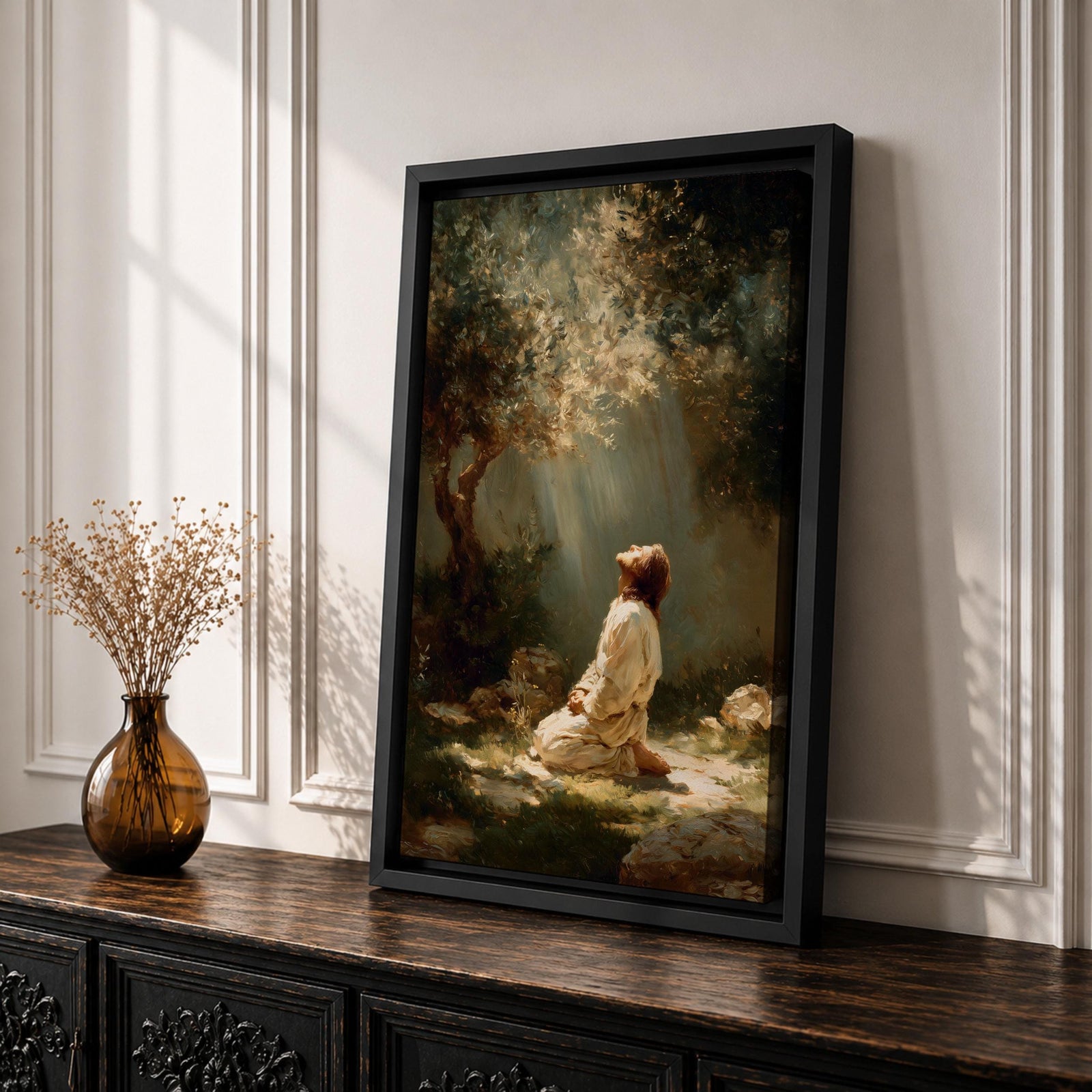 Framed artwork of Jesus praying in the Garden of Gethsemane, kneeling beneath a tree as soft rays of light shine down from above. Set in a modern living room, the canvas rests on a wooden console beside plants and neutral decor.