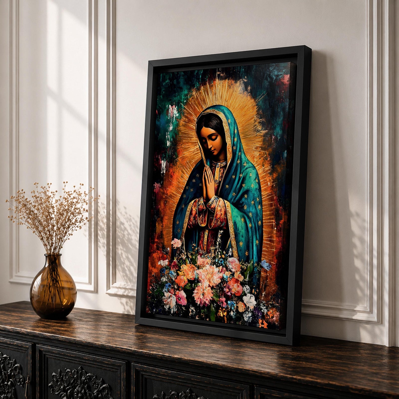 A vibrant framed canvas painting of Our Lady of Guadalupe depicts the Virgin of Guadalupe with hands clasped in prayer, wearing a teal mantle adorned with gold stars and surrounded by radiant light. The foreground features a colorful array of flowers, emphasizing the Catholic religious theme.