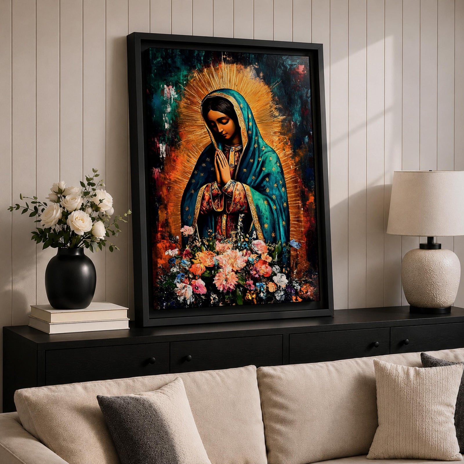 A vibrant framed canvas painting of Our Lady of Guadalupe depicts the Virgin of Guadalupe with hands clasped in prayer, wearing a teal mantle adorned with gold stars and surrounded by radiant light. The foreground features a colorful array of flowers, emphasizing the Catholic religious theme.