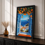 A vibrant framed canvas painting depicting an open Mediterranean-style window with blue shutters overlooking a coastal landscape. Above the window, ripe orange fruits hang from branches, framing a scenic view of white houses with terracotta roofs and a deep blue sea under a clear sky, rendered in a vivid, realistic style.
