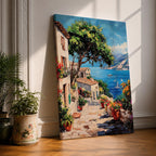This artwork depicts a vibrant Italian village scene with terracotta-roofed houses, lush greenery, and colorful flowers lining a stone pathway that leads down to a bright blue Mediterranean sea with sailboats and distant mountains, rendered in a realistic style capturing coastal charm.