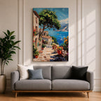 This artwork depicts a vibrant Italian village scene with terracotta-roofed houses, lush greenery, and colorful flowers lining a stone pathway that leads down to a bright blue Mediterranean sea with sailboats and distant mountains, rendered in a realistic style capturing coastal charm.