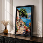 This artwork depicts a vibrant Italian village scene with terracotta-roofed houses, lush greenery, and colorful flowers lining a stone pathway that leads down to a bright blue Mediterranean sea with sailboats and distant mountains, rendered in a realistic style capturing coastal charm.