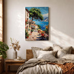 This artwork depicts a vibrant Italian village scene with terracotta-roofed houses, lush greenery, and colorful flowers lining a stone pathway that leads down to a bright blue Mediterranean sea with sailboats and distant mountains, rendered in a realistic style capturing coastal charm.