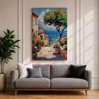 A vibrant framed canvas painting depicting a charming coastal village with stone-paved streets, terracotta rooftops, lush greenery, colorful flowers, and sailboats on a deep blue sea under a clear sky, rendered in a detailed, realistic style capturing Mediterranean summer ambiance.