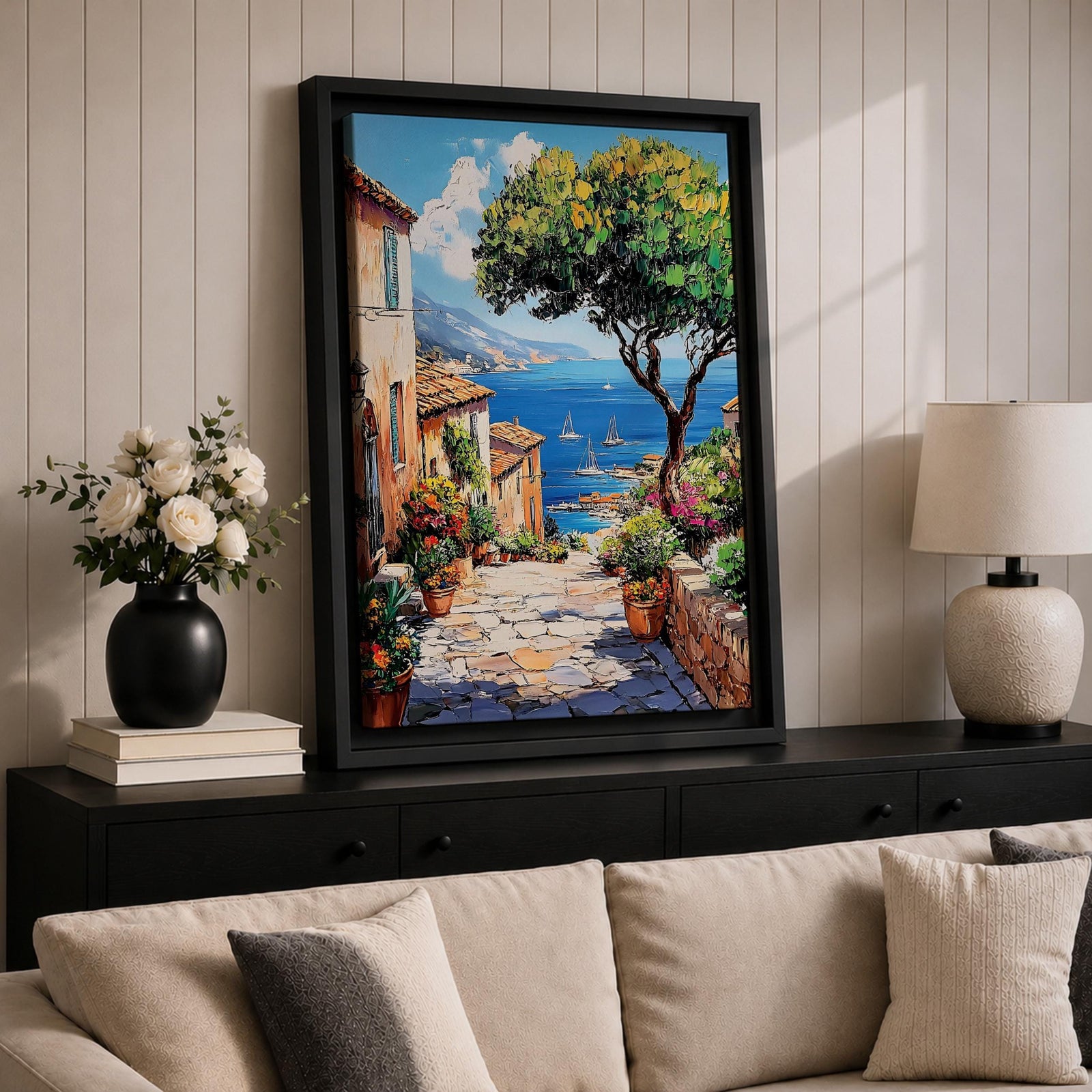 A vibrant framed canvas painting depicting a charming coastal village with stone-paved streets, terracotta rooftops, lush greenery, colorful flowers, and sailboats on a deep blue sea under a clear sky, rendered in a detailed, realistic style capturing Mediterranean summer ambiance.