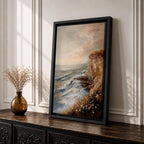 A vintage style framed canvas painting depicting a cliffside ocean seascape with waves crashing against rocky shores and wildflowers in the foreground. The textured brushwork captures the coastal landscape atmosphere with muted tones and a soft sky blending into the horizon.