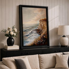A vintage style framed canvas painting depicting a cliffside ocean seascape with waves crashing against rocky shores and wildflowers in the foreground. The textured brushwork captures the coastal landscape atmosphere with muted tones and a soft sky blending into the horizon.