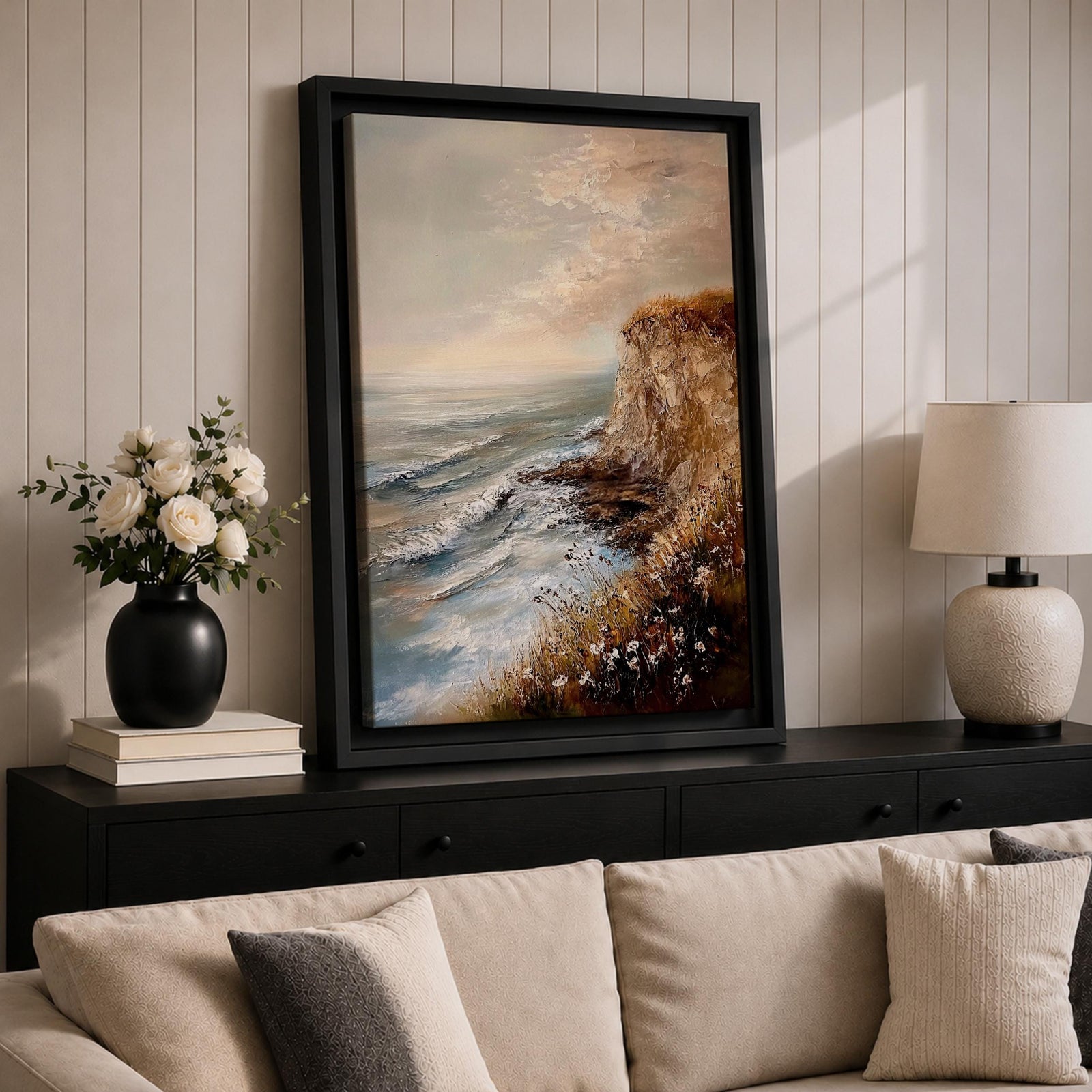 A vintage style framed canvas painting depicting a cliffside ocean seascape with waves crashing against rocky shores and wildflowers in the foreground. The textured brushwork captures the coastal landscape atmosphere with muted tones and a soft sky blending into the horizon.