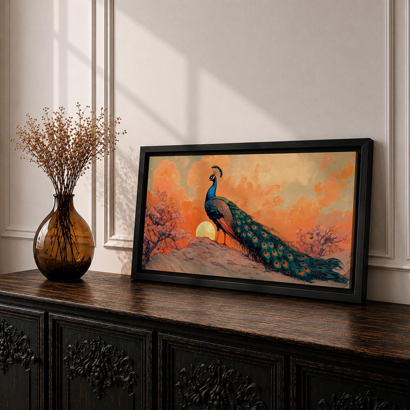 Panoramic artwork depicting a peacock standing on a rocky hill with its long iridescent tail feathers flowing behind it. The bird faces a glowing sunset with warm orange clouds and soft light illuminating the detailed blue and green plumage while flowering trees frame the scene.