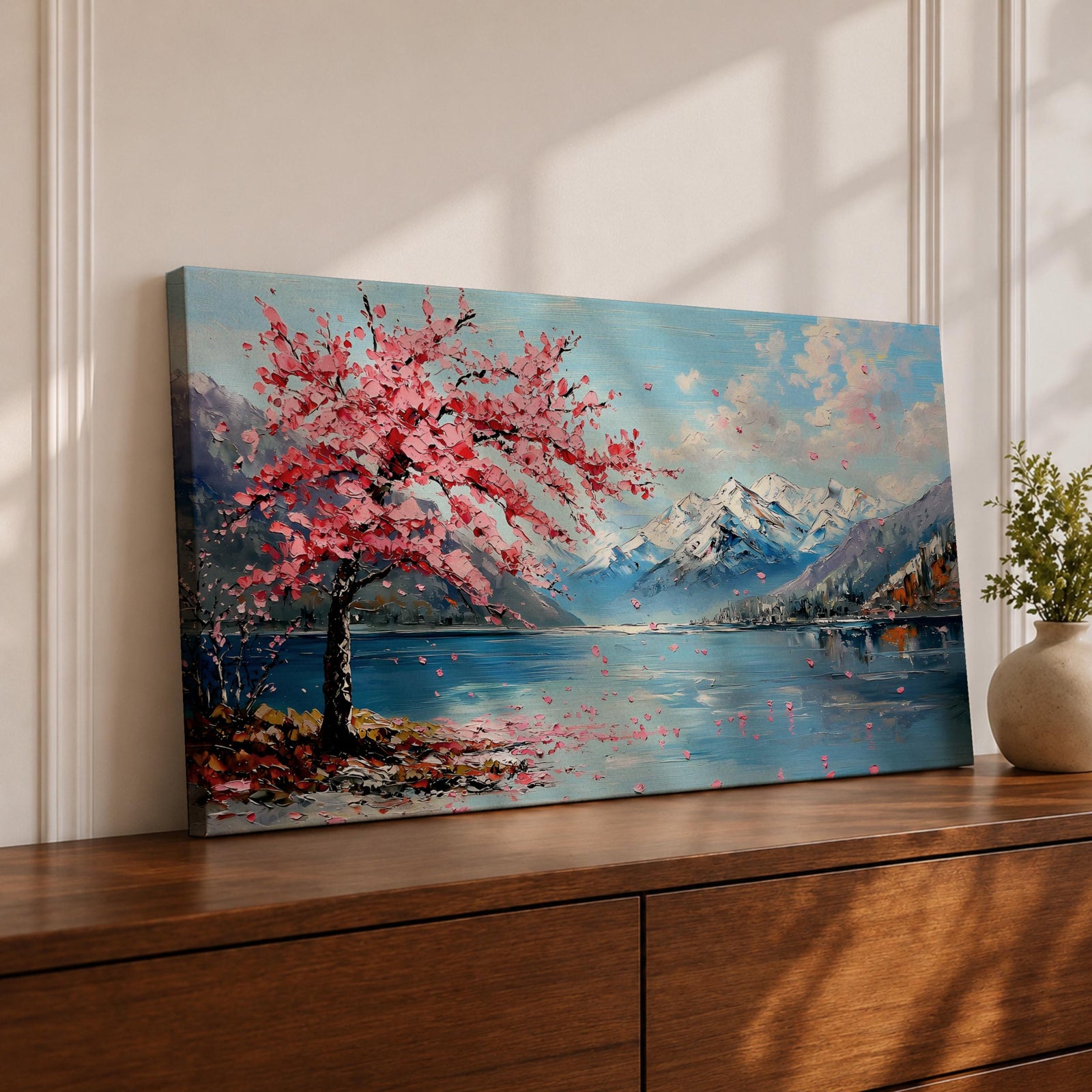 Panoramic painting of a cherry blossom tree in full bloom beside a calm mountain lake with snow covered peaks in the distance and soft clouds above. Pink petals drift across the water while thick palette knife brushstrokes create texture in the blossoms shoreline and reflections forming a wide spring landscape scene with bold color contrast.
