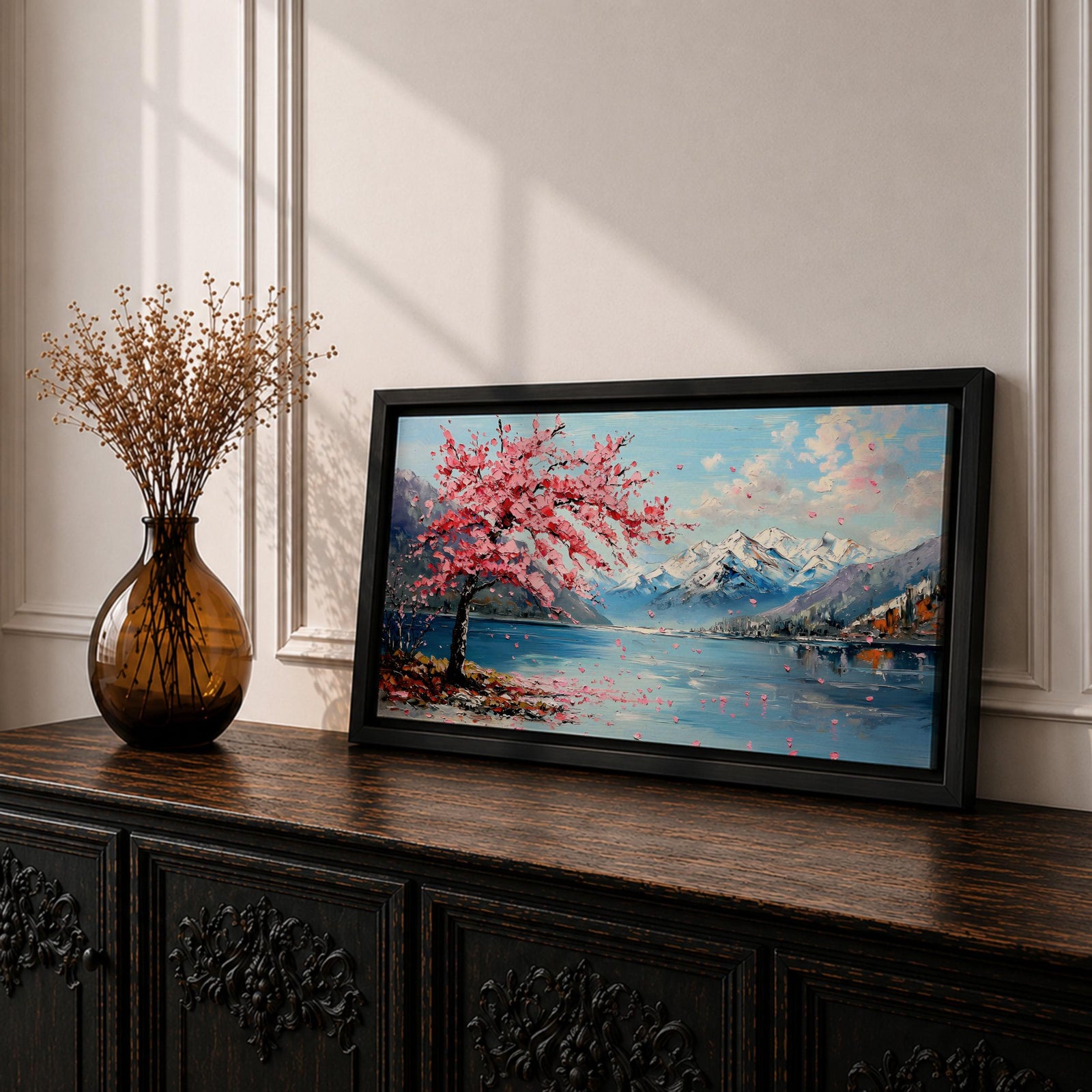 Panoramic painting of a cherry blossom tree in full bloom beside a calm mountain lake with snow covered peaks in the distance and soft clouds above. Pink petals drift across the water while thick palette knife brushstrokes create texture in the blossoms shoreline and reflections forming a wide spring landscape scene with bold color contrast.