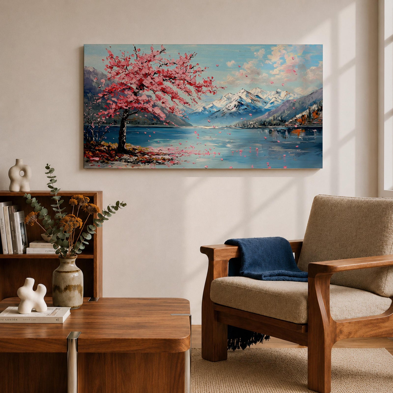Panoramic painting of a cherry blossom tree in full bloom beside a calm mountain lake with snow covered peaks in the distance and soft clouds above. Pink petals drift across the water while thick palette knife brushstrokes create texture in the blossoms shoreline and reflections forming a wide spring landscape scene with bold color contrast.