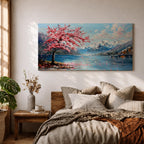 Panoramic painting of a cherry blossom tree in full bloom beside a calm mountain lake with snow covered peaks in the distance and soft clouds above. Pink petals drift across the water while thick palette knife brushstrokes create texture in the blossoms shoreline and reflections forming a wide spring landscape scene with bold color contrast.