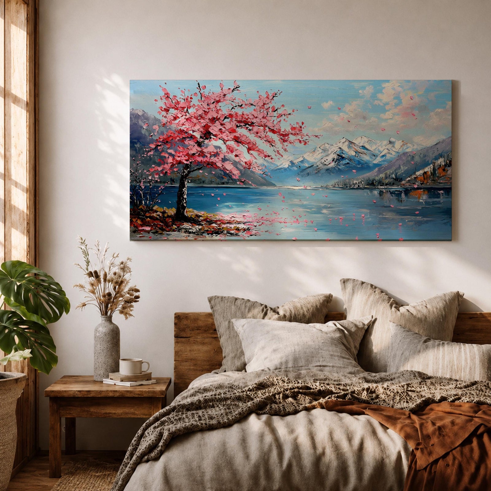 Panoramic painting of a cherry blossom tree in full bloom beside a calm mountain lake with snow covered peaks in the distance and soft clouds above. Pink petals drift across the water while thick palette knife brushstrokes create texture in the blossoms shoreline and reflections forming a wide spring landscape scene with bold color contrast.