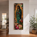A tall narrow vertical painting of the Virgin Mary Guadalupe shown in prayer with hands clasped, wearing a deep teal mantle adorned with gold stars and a soft pink dress, surrounded by radiant golden rays, with rich floral elements in vibrant pink, red, and cream tones at the base, rendered in a textured painterly style with warm, aged tones.