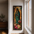 A tall narrow vertical painting of the Virgin Mary Guadalupe shown in prayer with hands clasped, wearing a deep teal mantle adorned with gold stars and a soft pink dress, surrounded by radiant golden rays, with rich floral elements in vibrant pink, red, and cream tones at the base, rendered in a textured painterly style with warm, aged tones.