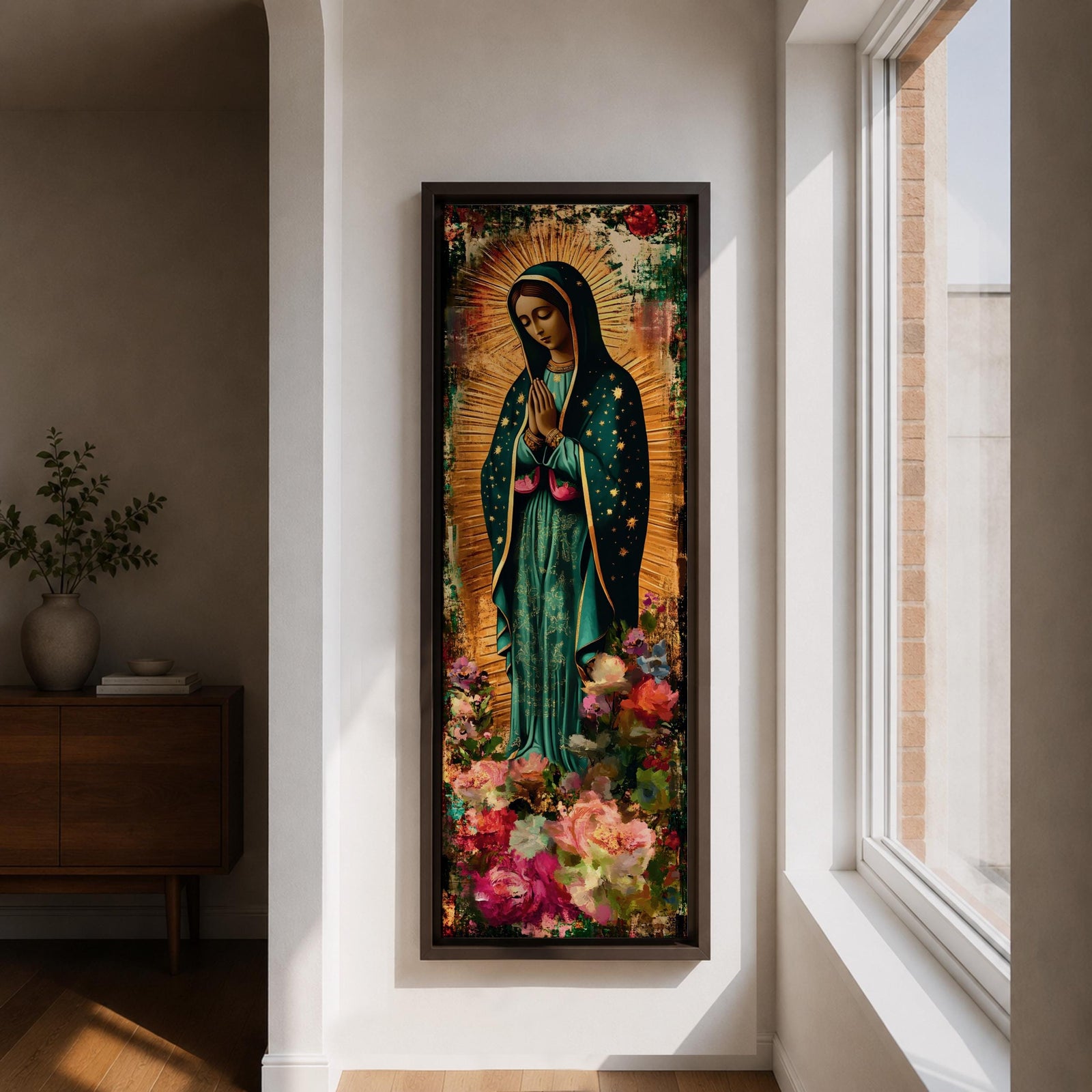 A tall narrow vertical painting of the Virgin Mary Guadalupe shown in prayer with hands clasped, wearing a deep teal mantle adorned with gold stars and a soft pink dress, surrounded by radiant golden rays, with rich floral elements in vibrant pink, red, and cream tones at the base, rendered in a textured painterly style with warm, aged tones.