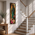 A tall narrow vertical painting of the Virgin Mary Guadalupe shown in prayer with hands clasped, wearing a deep teal mantle adorned with gold stars and a soft pink dress, surrounded by radiant golden rays, with rich floral elements in vibrant pink, red, and cream tones at the base, rendered in a textured painterly style with warm, aged tones.
