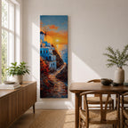 A tall vertical painting depicts a Santorini coastal scene with whitewashed buildings perched along a cliffside, a winding stone path leading toward the sea, and a glowing sunset casting warm orange and gold light across the sky and water, with a windmill silhouette and small sailboats adding depth to the vibrant Mediterranean composition.