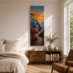 A tall vertical painting depicts a Santorini coastal scene with whitewashed buildings perched along a cliffside, a winding stone path leading toward the sea, and a glowing sunset casting warm orange and gold light across the sky and water, with a windmill silhouette and small sailboats adding depth to the vibrant Mediterranean composition.
