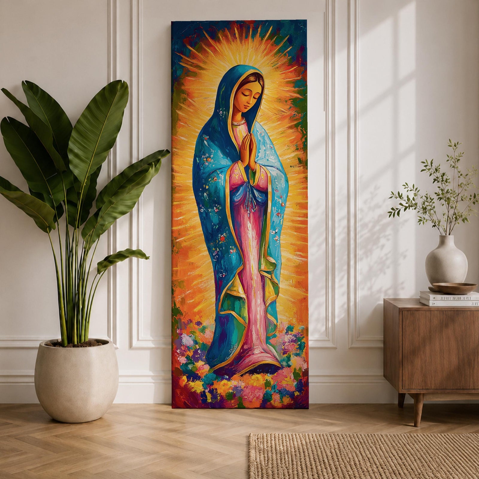 Long vertical painting of the Virgin Mary standing in prayer with hands together, wearing a blue mantle over a pink robe and surrounded by radiant golden rays. Colorful flowers gather at her feet while expressive brushstrokes and vibrant tones create a devotional depiction inspired by the Virgen de Guadalupe iconography.