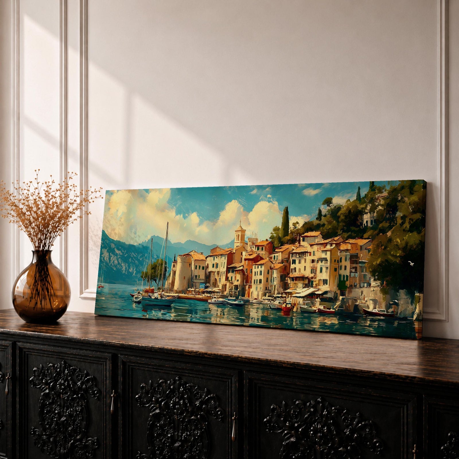 Panoramic coastal village painting showing colorful Mediterranean houses clustered along a harbor with sailboats resting on calm blue water. A small church tower rises above the waterfront while mountains and lush hills frame the background beneath a bright sky with soft clouds across the wide horizontal scene.