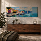 Panoramic Mediterranean harbor village painting featuring colorful seaside houses along a stone waterfront with small fishing boats resting in calm blue water. Soft clouds drift above distant cliffs and green hills while textured brushstrokes capture reflections and gentle movement across the wide coastal scene.