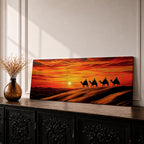 Panoramic desert sunset painting featuring a silhouette caravan of camels and riders walking along rolling sand dunes beneath a glowing red and orange sky. The low sun sits near the horizon while layered desert dunes stretch into the distance, creating a dramatic wide composition across the desert landscape.