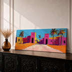 Panoramic desert village painting showing colorful Moroccan kasbah buildings in bright pink and warm ochre tones with tall palm trees lining a sandy road. Bold textured brushstrokes and vibrant colors create a lively North African scene beneath a clear blue sky across the wide horizontal composition.