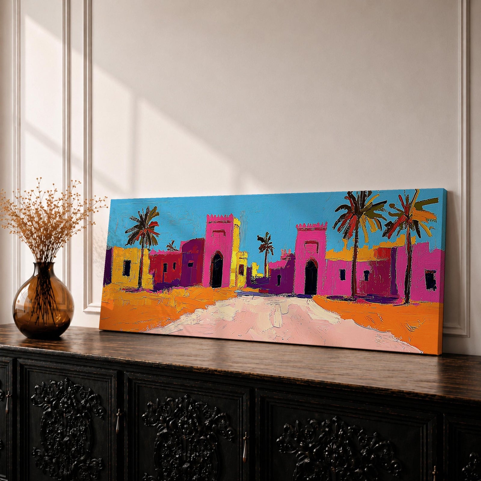Panoramic desert village painting showing colorful Moroccan kasbah buildings in bright pink and warm ochre tones with tall palm trees lining a sandy road. Bold textured brushstrokes and vibrant colors create a lively North African scene beneath a clear blue sky across the wide horizontal composition.