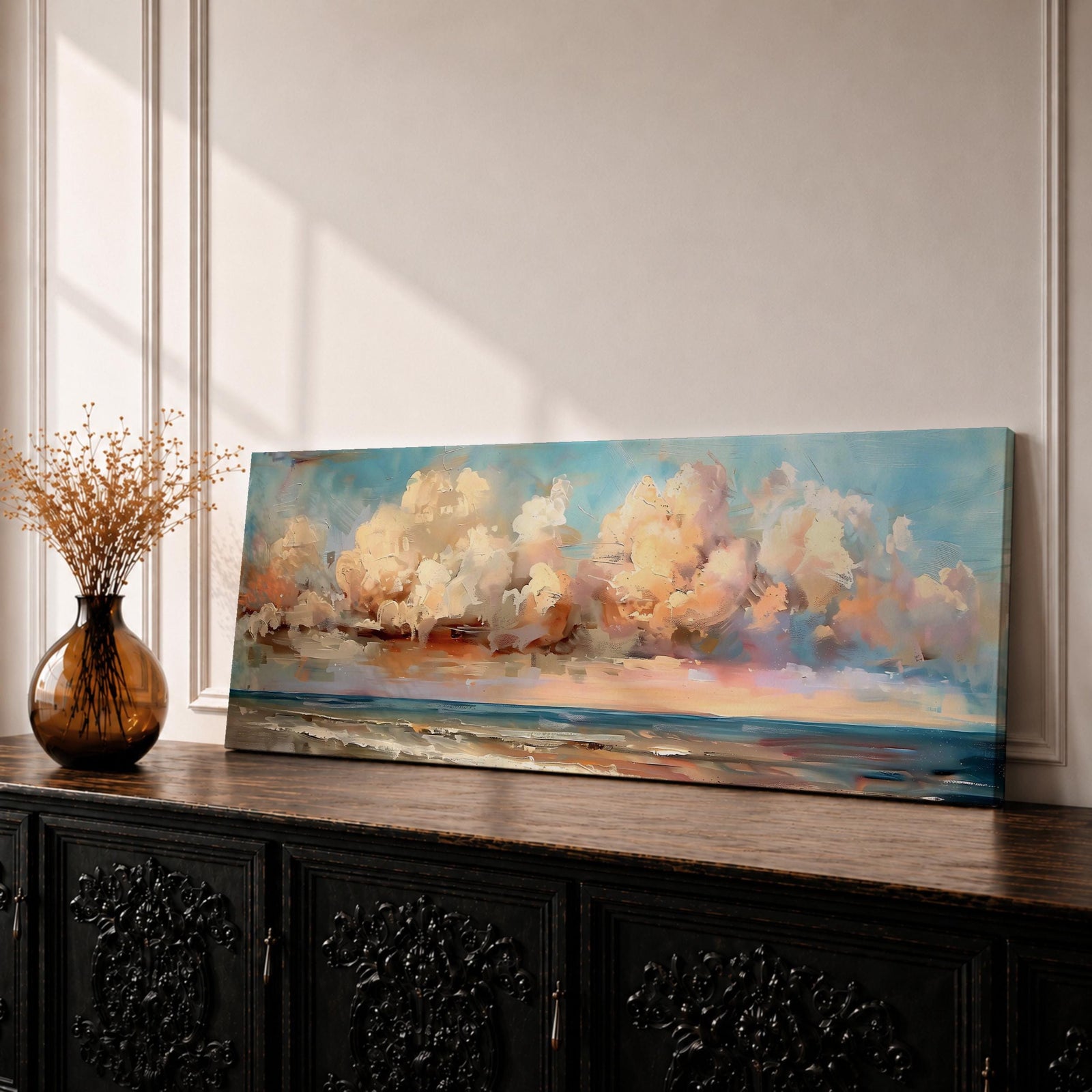 Panoramic coastal seascape painting of ocean waves reaching a sandy shoreline beneath expansive billowing clouds glowing with warm sunset tones. Soft peach, cream, and blue hues blend across the sky while gentle reflections shimmer along the water in a wide horizontal seaside composition.