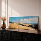 Panoramic wheat field landscape painting showing rolling golden farmland stretching across hills beneath a bright blue sky with scattered clouds. Textured brushstrokes depict tall wheat grasses in the foreground leading toward layered countryside fields, tree clusters, and distant hills across the wide rural horizon.