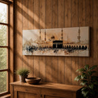 Panoramic painting of the Kaaba at Masjid Al Haram in Mecca surrounded by mosque arches and tall minarets. The sacred black cube stands at the center of a wide courtyard with crowds gathered around while warm golden tones and architectural details stretch across the expansive skyline.