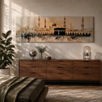 Panoramic painting of the Kaaba at Masjid Al Haram in Mecca surrounded by mosque arches and tall minarets. The sacred black cube stands at the center of a wide courtyard with crowds gathered around while warm golden tones and architectural details stretch across the expansive skyline.