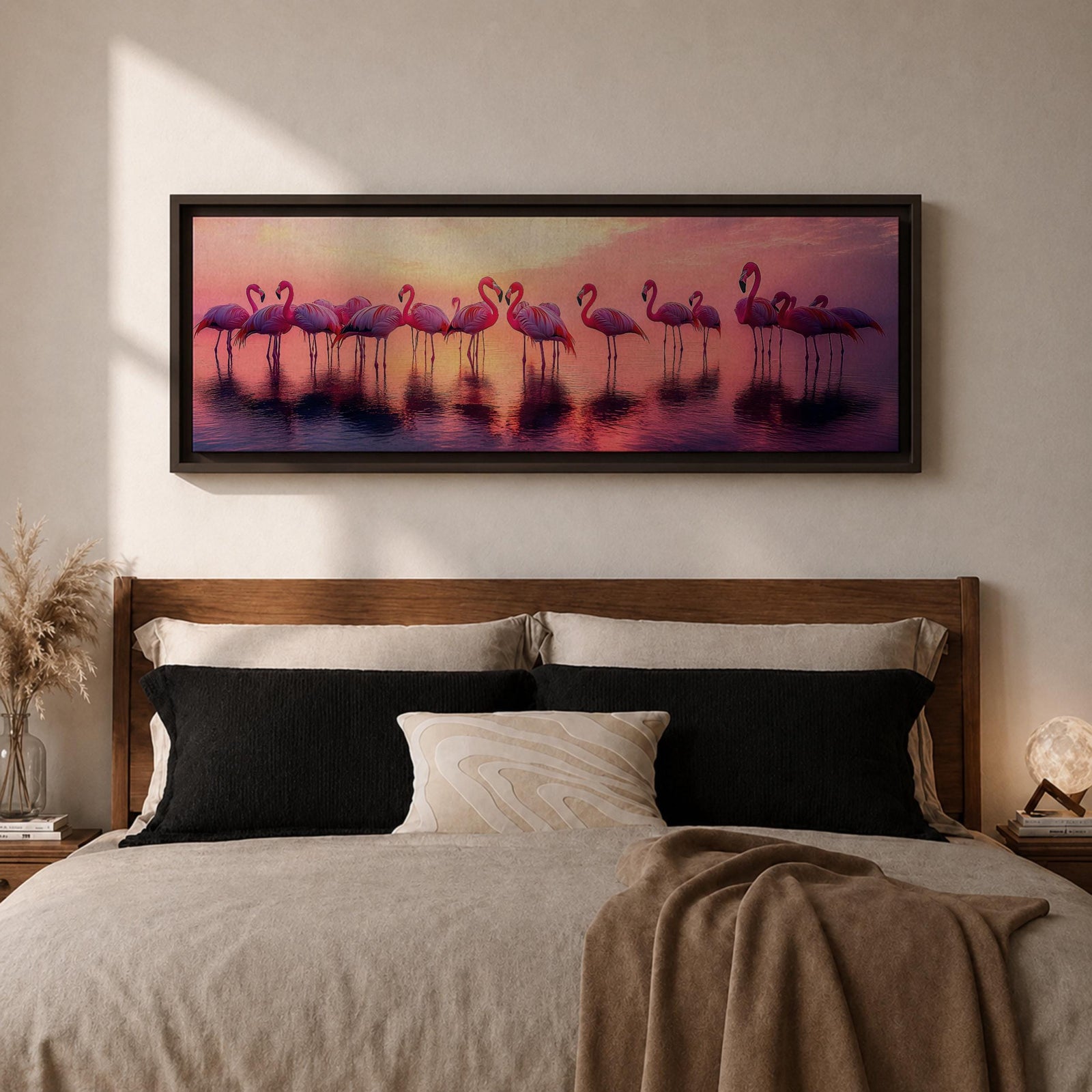 Panoramic artwork showing a flock of flamingos standing in shallow reflective water at sunset. The long horizontal composition features pink birds mirrored in the calm lake surface with glowing orange, purple, and magenta sky tones behind them, creating a tropical waterscape with silhouettes and soft reflections stretching across the scene.