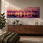 Panoramic artwork showing a flock of flamingos standing in shallow reflective water at sunset. The long horizontal composition features pink birds mirrored in the calm lake surface with glowing orange, purple, and magenta sky tones behind them, creating a tropical waterscape with silhouettes and soft reflections stretching across the scene.