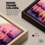 Panoramic artwork showing a flock of flamingos standing in shallow reflective water at sunset. The long horizontal composition features pink birds mirrored in the calm lake surface with glowing orange, purple, and magenta sky tones behind them, creating a tropical waterscape with silhouettes and soft reflections stretching across the scene.