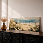 A long panoramic coastal painting showing sandy dunes covered with wild grasses and small white flowers overlooking a calm ocean shoreline. Soft waves roll toward the beach beneath a pale blue sky while textured brush strokes create a peaceful seascape landscape stretching across a wide horizontal composition.