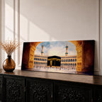 A long panoramic artwork depicting the Kaaba at the center of Masjid Al Haram in Mecca framed by grand mosque arches and tall minarets. The sacred black cube stands in the courtyard while pilgrims gather around it, creating a wide horizontal composition highlighting the holy Islamic sanctuary.