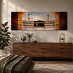 A long panoramic artwork depicting the Kaaba at the center of Masjid Al Haram in Mecca framed by grand mosque arches and tall minarets. The sacred black cube stands in the courtyard while pilgrims gather around it, creating a wide horizontal composition highlighting the holy Islamic sanctuary.