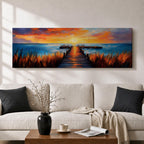 A long narrow panoramic painting depicts a wooden pier extending through tall grasses into calm ocean waters, leading toward a glowing sunset on the horizon, with bold textured brushstrokes and vivid color contrasts capturing reflections of warm light across the water and sky in an expressive contemporary style.